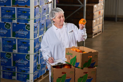 Women in white lab coat inspecting an orange bell pepper in produce cooler