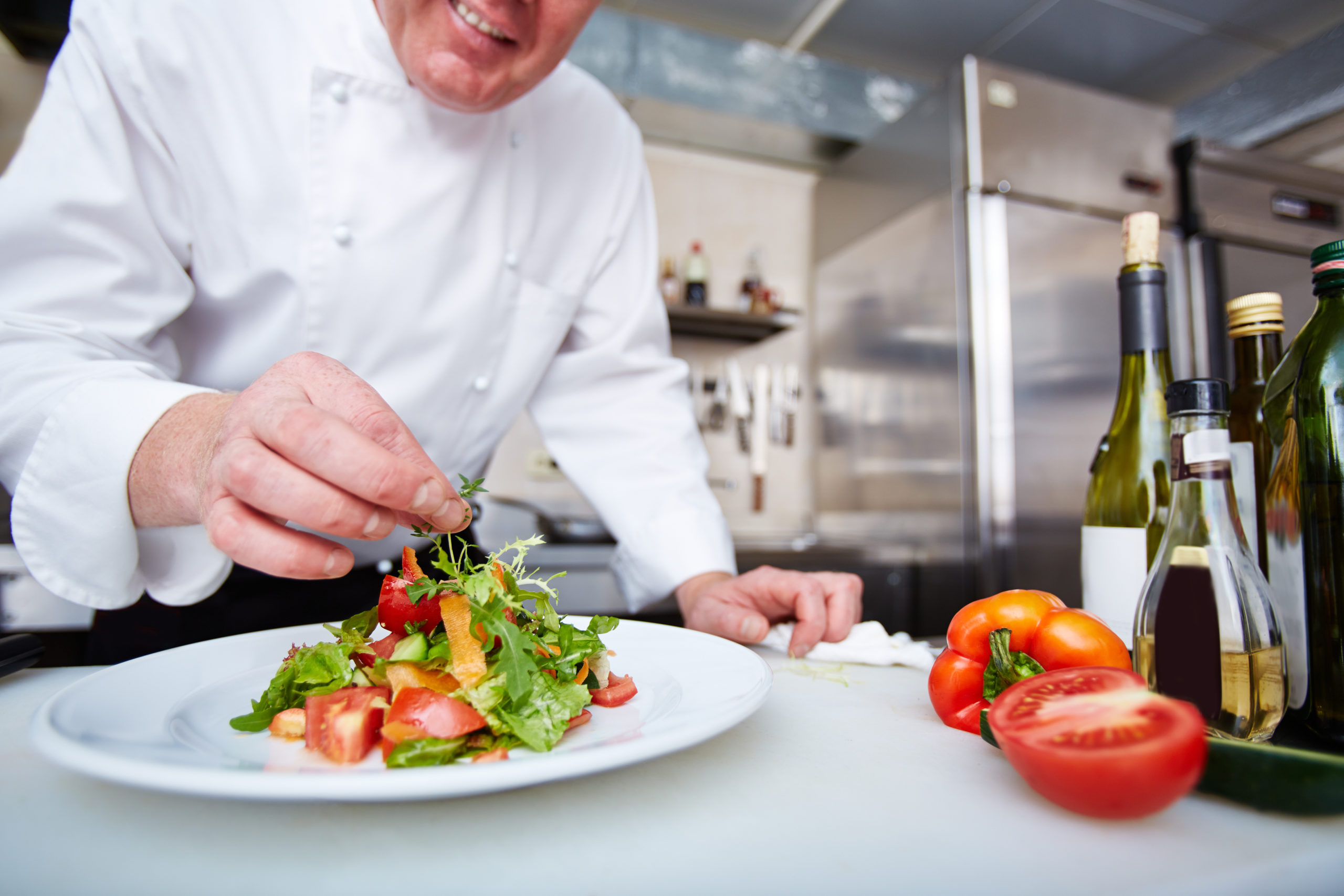 A man in a white chef coat adding herbs to a plate of salad